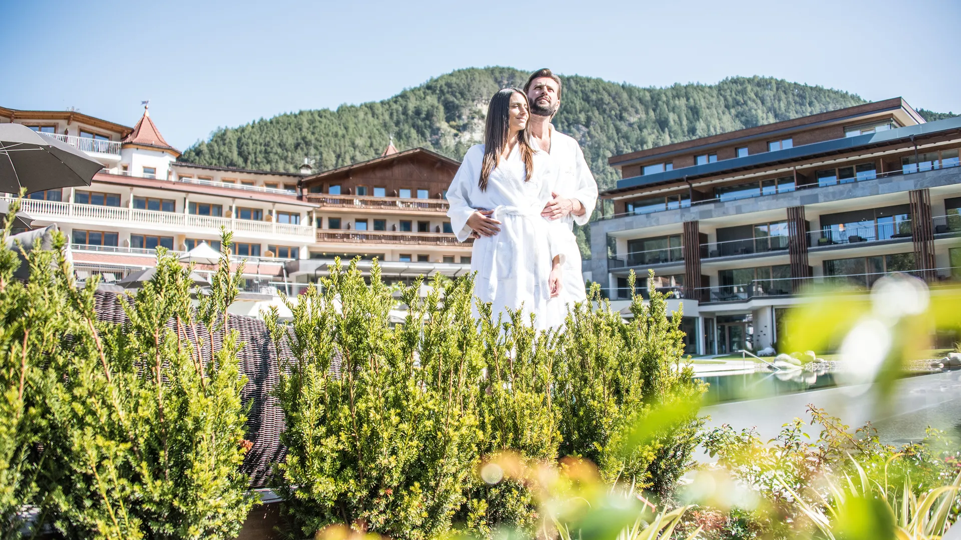 Paar im Garten vor Hotelgebäude mit Südtiroler Architektur und bewaldeten Bergen in Kiens
